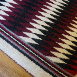 Square flatwoven rug featuring a burgundy, black, and cream serrated diamond pattern on hardwood flooring.