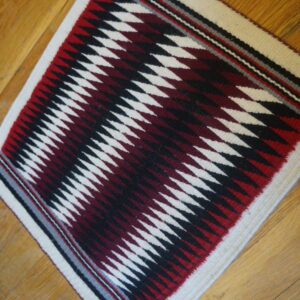 Geometric square flatwoven rug featuring white, black, and deep red zigzagging diamonds on wood planks.