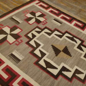 Low-pile rug with geometric patterns in gray-brown, dark brown, cream, and red on textured wood planks.