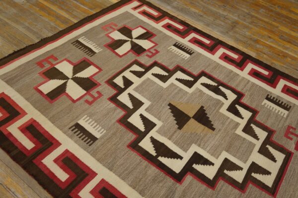 Low-pile rug with geometric patterns in gray-brown, dark brown, cream, and red on textured wood planks.