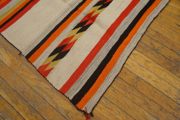 Cream flatweave rug with red, orange, black, and gold stripes, including a central zigzag pattern, on wood planks.