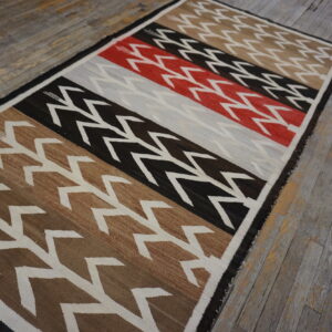 Low-pile rug with geometric zigzags across wide bands of brown, black, red, and gray on distressed wood floors.