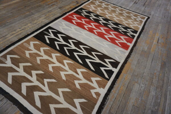 Low-pile rug with geometric zigzags across wide bands of brown, black, red, and gray on distressed wood floors.