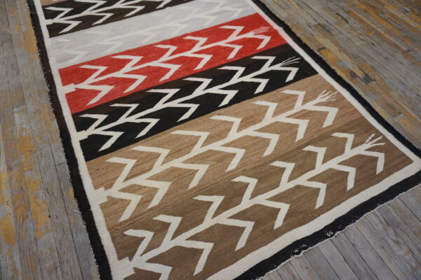 Geometric flatweave rug in red, black, tan, and gray colors with white zigzag motifs on wood planks.