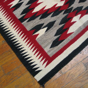 Flatweave rug corner with red, black, cream, and gray zigzag patterns sits on hardwood planks.