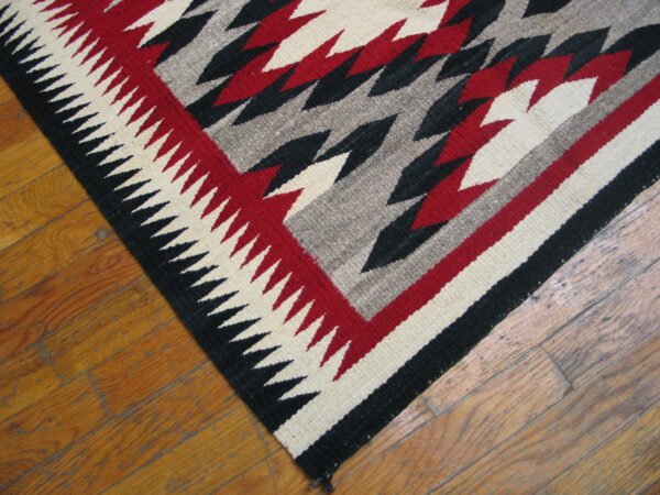 Flatweave rug corner with red, black, cream, and gray zigzag patterns sits on hardwood planks.