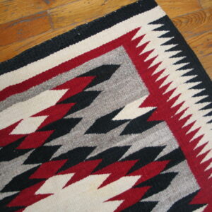 Geometric flatweave rug corner featuring black, red, cream, and gray zig-zags on wood plank flooring.