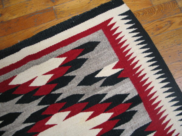 Geometric flatweave rug corner featuring black, red, cream, and gray zig-zags on wood plank flooring.