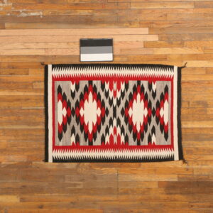 A flatweave rug with red, black, white, and gray geometric patterns rests on wooden plank flooring.