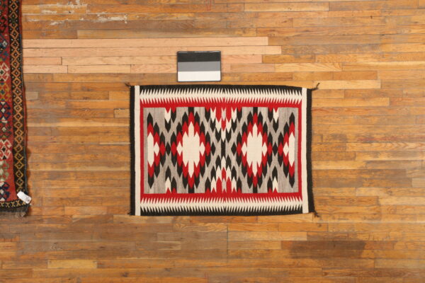 A flatweave rug with red, black, white, and gray geometric patterns rests on wooden plank flooring.