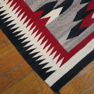 A flatwoven rug with black, red, gray, and cream serrated geometric patterns rests on a hardwood floor.