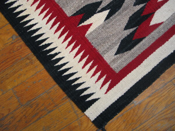 A flatwoven rug with black, red, gray, and cream serrated geometric patterns rests on a hardwood floor.