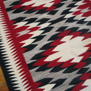 Geometric low-pile rug featuring serrated red, black, and white diamonds on a grey field over a wood floor.