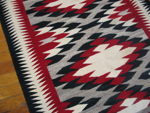 Geometric low-pile rug featuring serrated red, black, and white diamonds on a grey field over a wood floor.