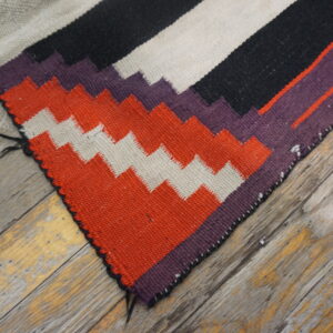 A flatweave rug featuring black, beige, orange, and purple geometric patterns rests on distressed wood flooring.