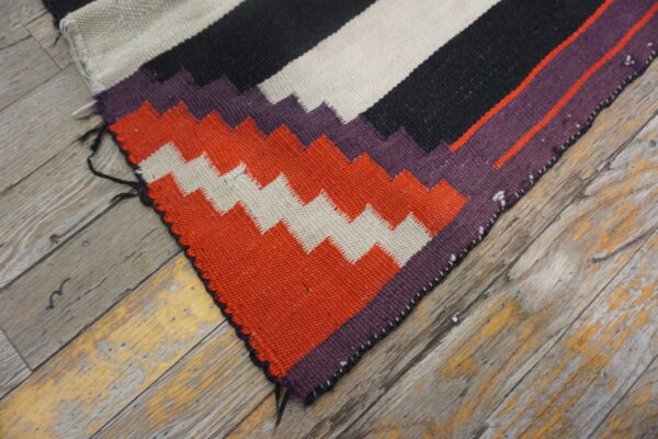 A flatweave rug featuring black, beige, orange, and purple geometric patterns rests on distressed wood flooring.