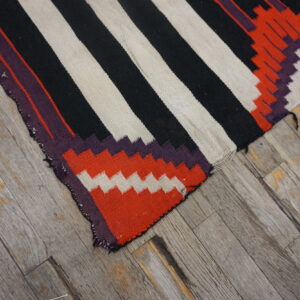 Close-up of a striped black, off-white, and geometric red flatweave rug on aged wood plank flooring.
