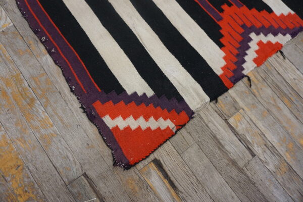 Close-up of a striped black, off-white, and geometric red flatweave rug on aged wood plank flooring.