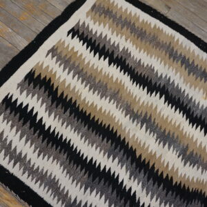 Flatweave rug featuring diagonal zigzag stripes in black, cream, tan, and heathered gray on aged wooden floorboards.