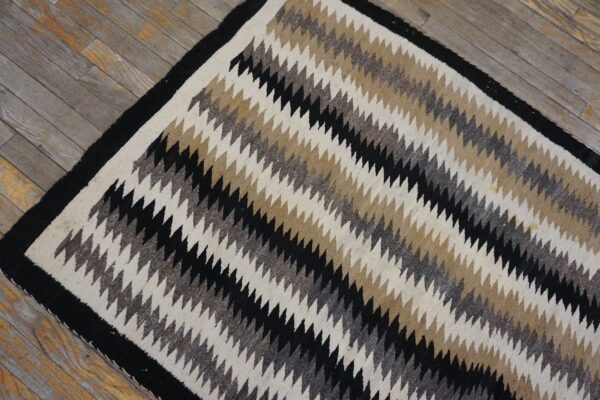 Flatweave rug featuring diagonal zigzag stripes in black, cream, tan, and heathered gray on aged wooden floorboards.
