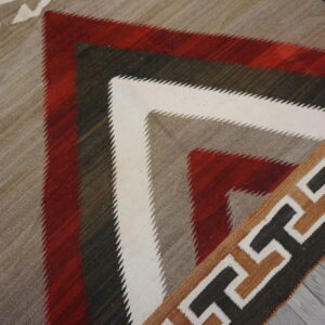 Flatweave rug with serrated geometric patterns in red, white, gray, and brown, resting on gray wood flooring.