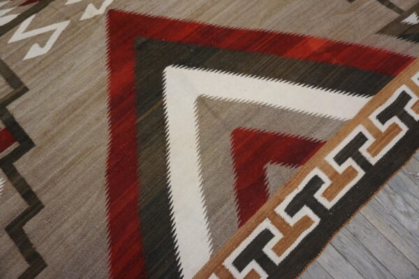 Flatweave rug with serrated geometric patterns in red, white, gray, and brown, resting on gray wood flooring.