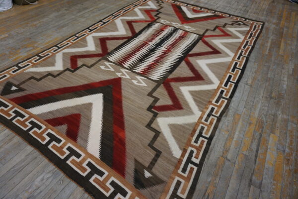 Geometric flat-weave rug in tan, red, white, and dark brown on a distressed wood floor.