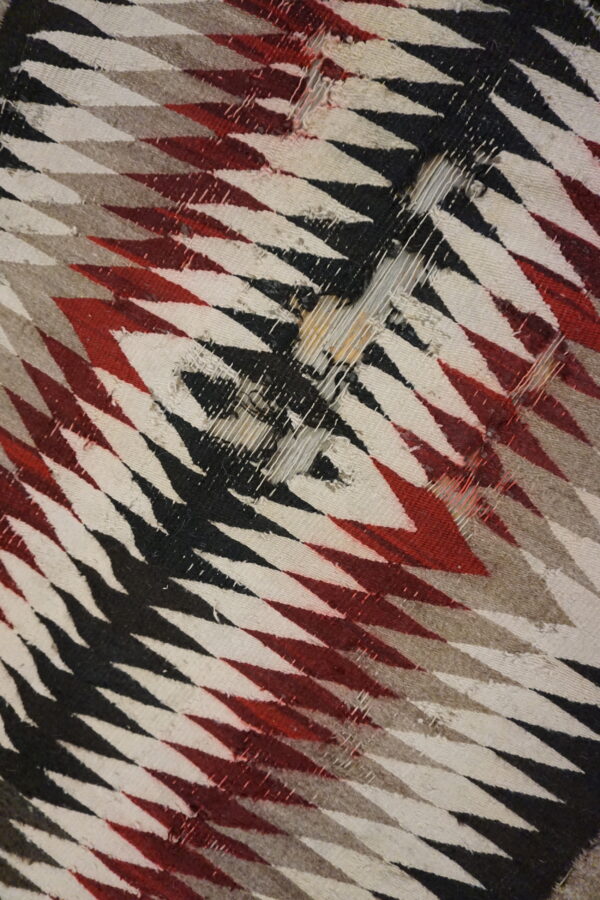 Worn flatweave rug detail with red, black, cream, and taupe diagonal zigzag patterns.