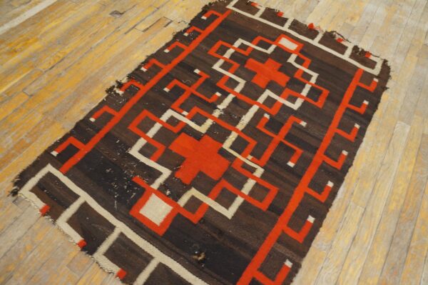 Dark brown flatweave rug featuring interlocking red and cream geometric designs on a distressed wood floor.