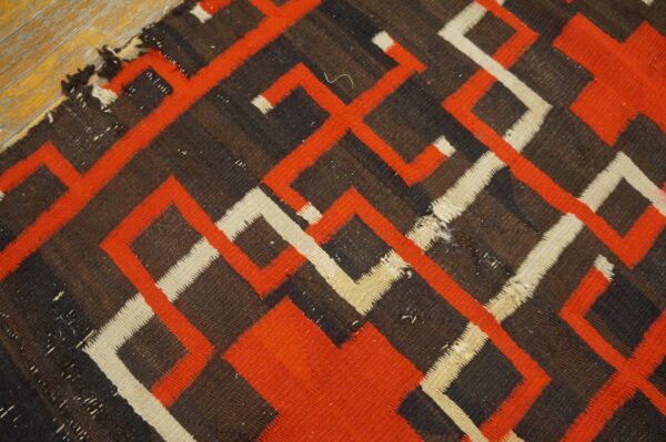 Worn flatweave rug with striking orange, cream, and brown geometric patterns lying on a wood floor.
