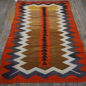 A flatwoven rug featuring orange, brown, black, white, and yellow chevron patterns lies on distressed wood flooring.