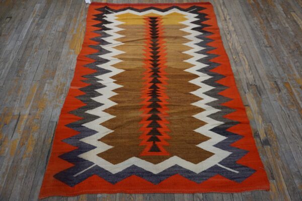 A flatwoven rug featuring orange, brown, black, white, and yellow chevron patterns lies on distressed wood flooring.
