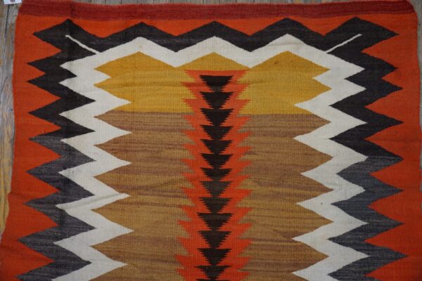 Low-pile rug with a central spine of dark triangles flanked by thick zigzags in orange, mustard, white, and black.