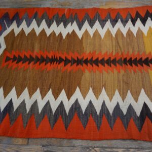 A brightly colored flatweave rug features large serrated zig-zag patterns in orange, brown, white, and dark gray.