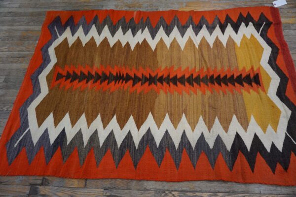 A brightly colored flatweave rug features large serrated zig-zag patterns in orange, brown, white, and dark gray.