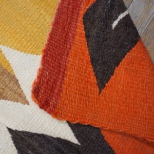 Close-up of a woven rug displaying yellow, orange, red, and dark brown geometric shapes on wooden floorboards.