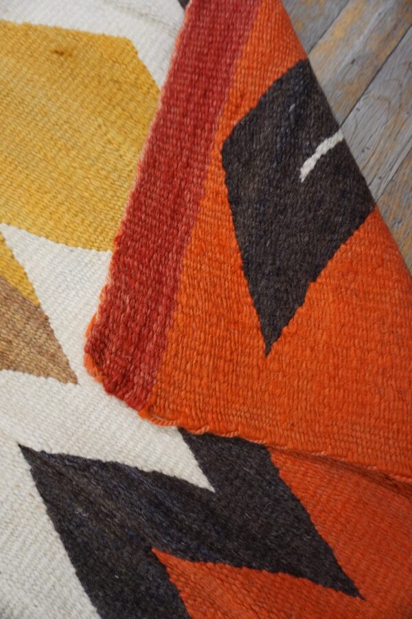 Close-up of a woven rug displaying yellow, orange, red, and dark brown geometric shapes on wooden floorboards.