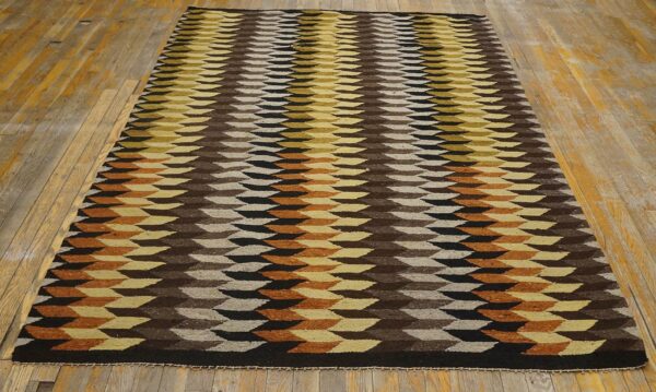 Geometric flatweave rug in brown, yellow, black, and rust repeating shapes displayed on worn wooden flooring.