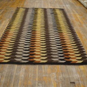 Flatweave rug featuring brown, yellow, black, and beige geometric blocks rests on worn wood flooring.