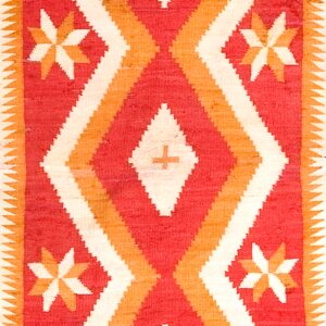 Geometric flatweave rug in red, cream, and gold with zigzag borders rests on distressed wood planks.