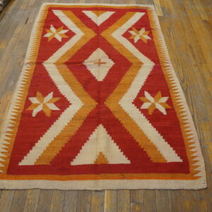 Red, orange, and cream flatweave rug with geometric patterns displayed on worn wood flooring.