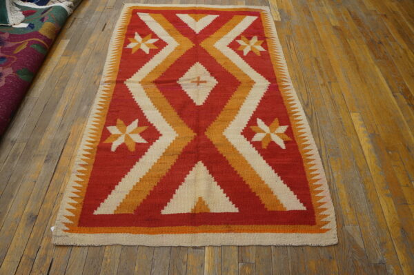Red, orange, and cream flatweave rug with geometric patterns displayed on worn wood flooring.