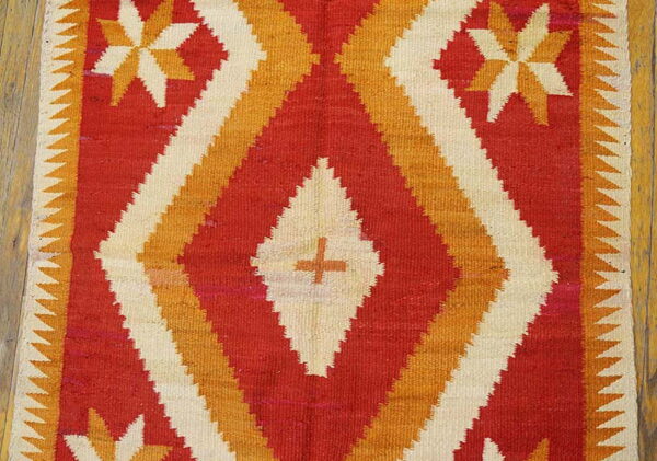 Flatweave rug in red, orange, and cream with stepped geometric patterns resting on wood flooring.