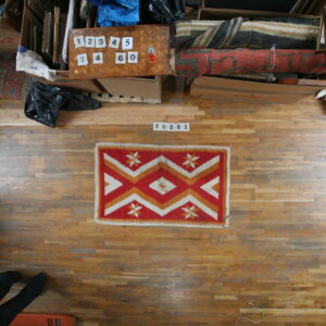 Geometric red, white, and orange rug displayed flat on distressed wood floorboards amongst storage items.