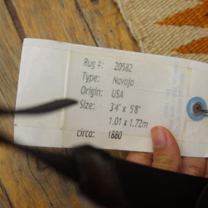 White informational tag is attached to a textured rug featuring cream, orange, and red geometric patterns on wood.