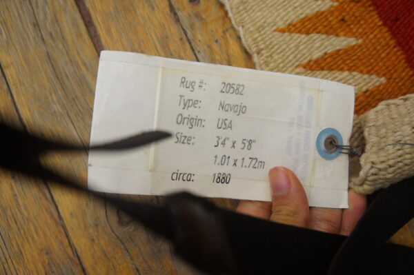 White informational tag is attached to a textured rug featuring cream, orange, and red geometric patterns on wood.