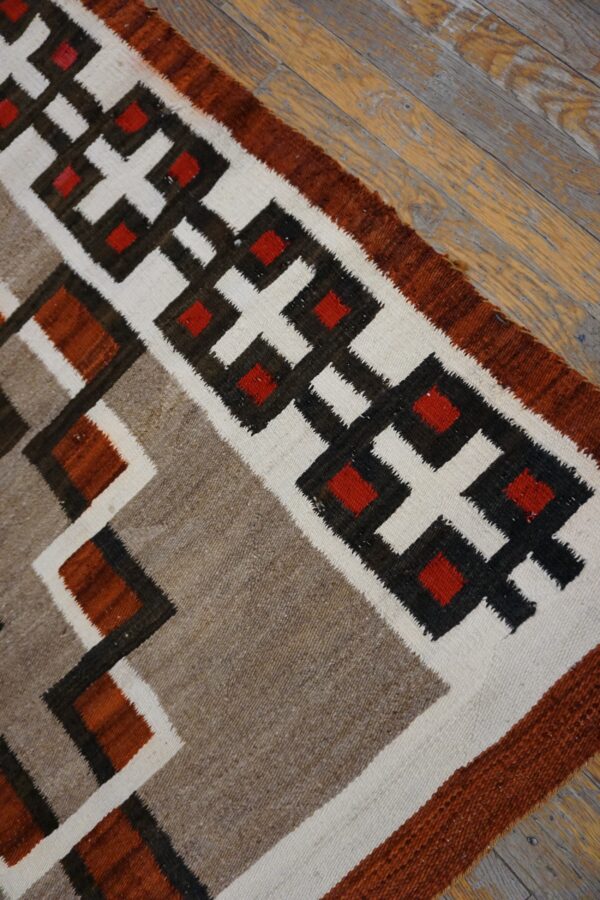 Geometric flatweave rug in red, brown, white, and tan featuring interlocking stepped patterns on distressed wood.