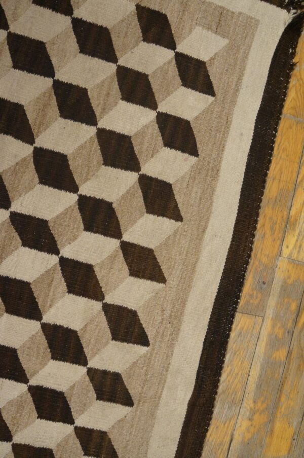 Dark brown and beige tumbling block patterned flatweave rug lies partially on worn wooden floorboards.