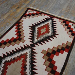 Geometric flatweave rug with serrated diamond motifs in white, red, dark brown, and tan on weathered planks.