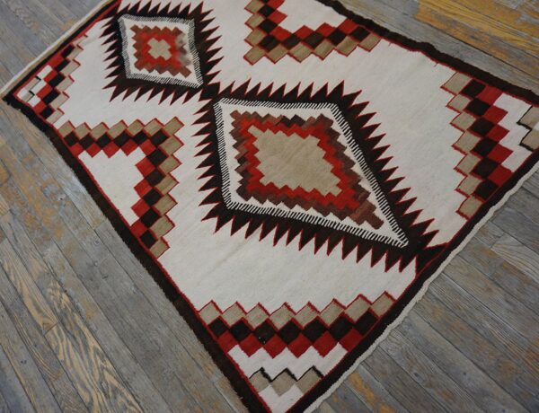 White flatweave rug with dark red, brown, and tan geometric diamond motifs rests on rustic gray wood flooring.